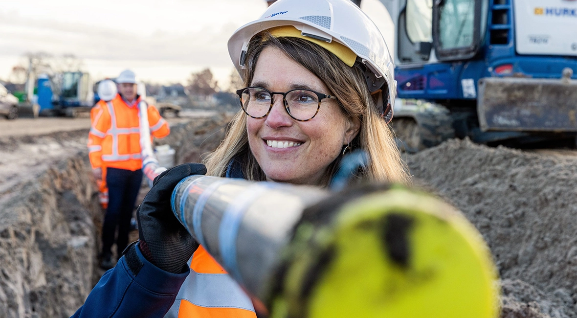 Lächelnde Frau in Arbeitskleidung hält zusammen mit einem Mann im Hintergrund ein Rohr (Foto)